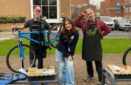 Three individuals pose for a photograph as they work on repairing a bicycle