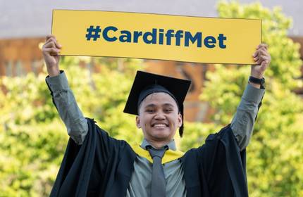 A Cardiff Met graduate in cap and gown holds a yellow #CardiffMet sign above their head