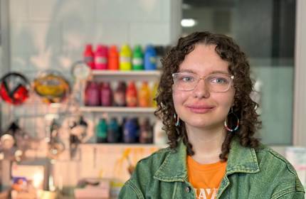 A person with curly hair and glasses smiles in a colourful art studio. They wear a denim jacket over an orange shirt. In the background, shelves display vibrant paint bottles and art supplies.