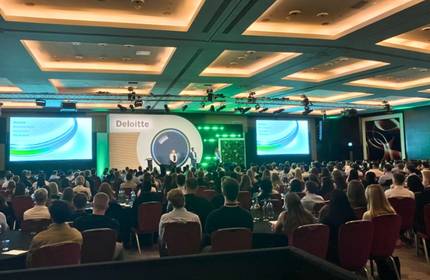A large conference room filled with seated attendees facing a stage. The stage has a speaker, green lighting, and a screen displaying the Deloitte logo and presentation slides. The ceiling features recessed lighting.