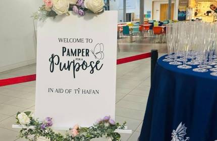 A sign with floral decorations reads Welcome to Pamper for a Purpose in aid of Ty Hafan. Beside it, a table holds numerous empty champagne flutes, and colourful chairs are visible in the background.