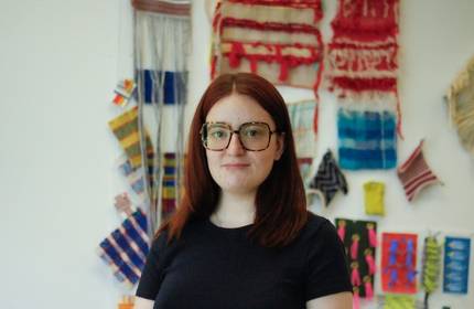 A woman with glasses stands in front of a colourful wall of textiles, showcasing various fabric patterns and textures.