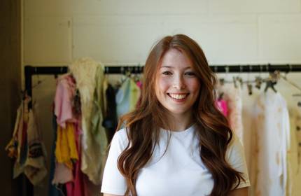 A woman with long auburn hair and a white shirt smiles at the camera, standing in front of a clothing rack with colourful garments.
