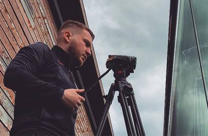 Jonathan Aveston, wearing a dark-coloured tracksuit, stands behind a tripod-mounted camcorder