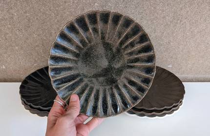 A hand holding a dark ceramic plate with a ribbed pattern. Several similar plates are stacked in the background on a white surface, against a neutral-coloured wall.