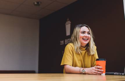 Kristine Clifford, sat at a wooden desk, holds an orange takeaway coffee cup