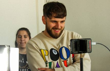 Man in a cream graphic sweatshirt looks into a digital camera set on a stand