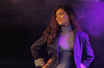 Nuwanee, wearing a textured grey turtleneck and black suit jacket, poses for a photograph under purple lighting