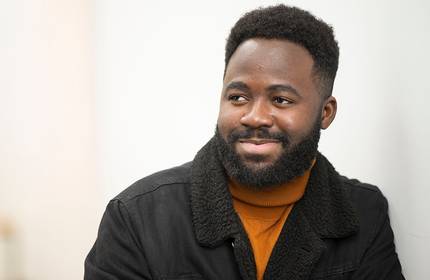 Ted Airaga, wearing an orange turtleneck and black jacket, leans against a white wall