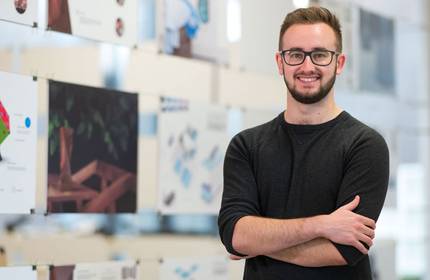 Tom Fantom stands with arms crossed in front of a glass display board containing various pictures and design illustrations