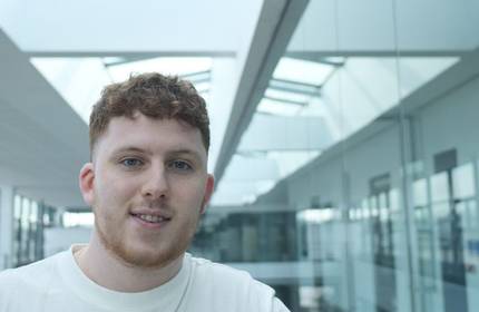 A young man with curly hair stands confidently in a well-lit hallway, showcasing a relaxed demeanor.