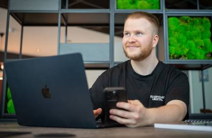 A man wearing a black shirt is seated at a table with a laptop and a cell phone in front of him.