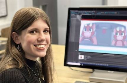 A person with long hair smiles at the camera while sitting at a desk. In the background, a computer screen displays an animation of two cartoon characters driving a car.