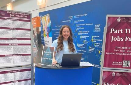 A young woman stands by a table displaying a sign for a part-time jobs fair, engaging with attendees.
