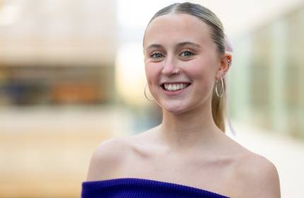 A young woman with hoop earrings and a nose ring smiles while wearing an off-the-shoulder blue top. The background is blurred, highlighting her cheerful expression.