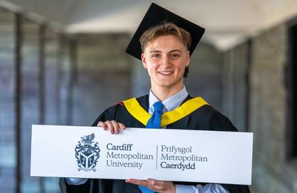 A Cardiff Met graduate in cap and gown holds a Cardiff Met-branded placard