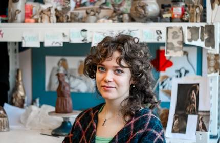 A person with curly hair sits in a creative workspace filled with art supplies and drawings. Shelves behind them display sculptures and sketches, adding to the artistic atmosphere. They wear a checkered shawl and a light green top.
