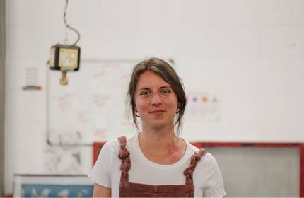A woman wearing an apron stands in front of a textured wall, smiling and looking towards the camera.