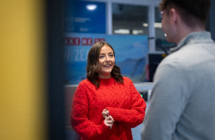 A young woman in a knitted red jumper talks to someone facing away from the camera