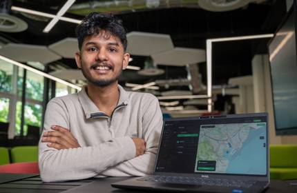 A young man sits at a desk, showing off their work on a laptop