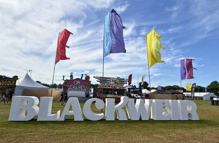 A large Blackweir sign under four colourful flags in front of an open air concert field