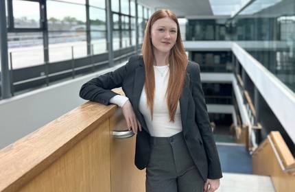 A young woman in casual grey business suit leans against a wooden support rail in a large, airy building