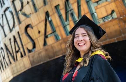 A Cardiff Met graduate in cap and gown stands in front of the Wales Millennium Centre