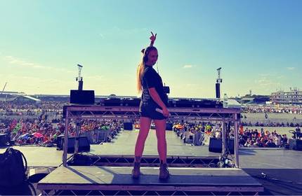 A DJ stands on an outdoor stage in front of a large, seated audience. The sky is clear and sunny. The DJ, with long hair, is making a peace sign with one hand. In the background, there are buildings and structures.