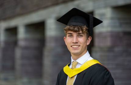 A young man in a graduation cap and gown stands smiling. He wears a white shirt, light brown tie, and has a yellow sash. The background is a stone wall in soft focus.