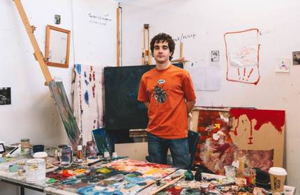 A young adult stands behind the desk in a messy art studio