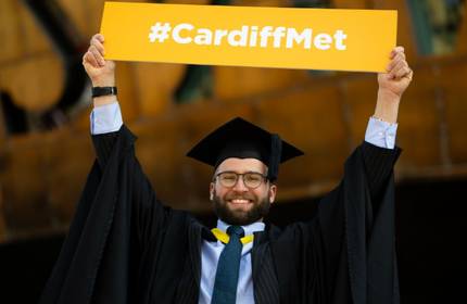 A graduate wearing a cap and gown joyfully holds up a yellow sign with #CardiffMet written on it.