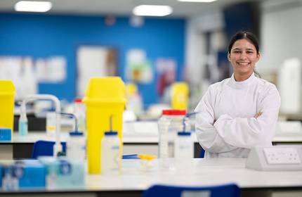A woman wearing a lab coat stands beside a table, prepared for scientific work or analysis in a laboratory setting.