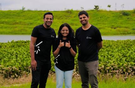 Three people in black shirts smile outdoors by a lake and greenery. The person in the middle gives a thumbs-up. Its a bright, slightly cloudy day, with a grassy hill in the background.