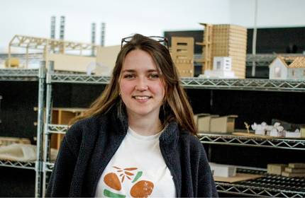 Young female wearing a white t-shirt and fleece jacket stands in front of metal shelves containing various small wooden building models