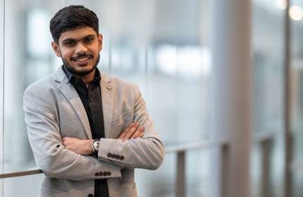 Jay Kiranbhai Patel, wearing a light gray suit and black shirt, stands in an open, airy building walkway