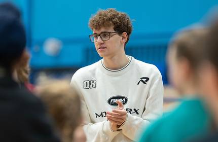 A young man with glasses and wearing a white sweatshirt clasps his hands together