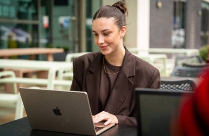 A young woman in a dark shirt and blazer works at a table on a laptop in an outdoor seating area