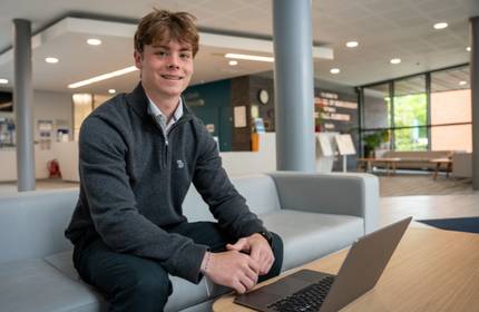 A young man in a dark jumper sits forward on a sofa looking at a laptop on a coffee table in front of them