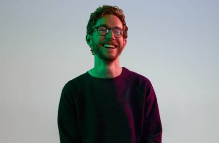 A young man wearing glasses smiles under low lighting