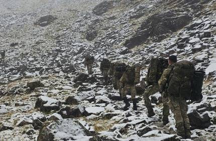 A group of soldiers in camouflage jumpsuits and carrying large backpacks hike in single file through a large, open hillside covered in slate and rocks