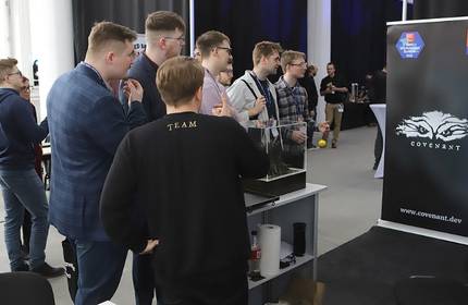 A group of people stands near a table with a computer setup, engaging in discussion. They are in an indoor event space with banners displaying the word Covenant and accolades like Great Place to Work in the background.