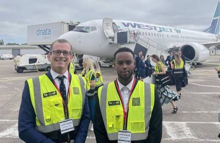 Two people wearing hi-vis vests stand on the tarmac of an airport. In the background are a group of people and a passenger airplane.