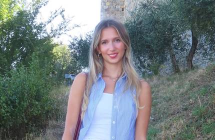 A young woman wearing a sleeveless blue shirt stands in front of some trees and an old stone building.