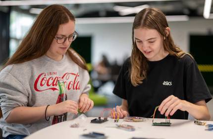 Two young women sit alongside each other at a table. On the table are a number of small electronic circuit boards with wires.