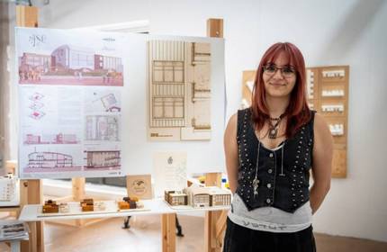 A young woman stands in front of a display of architectural drawings and small models of buildings.