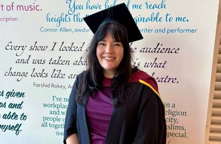 A woman wearing a graduation cap and gown stands in front of a wall on which a number of quotes have been printed.