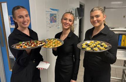 Three people stand in a professional kitchen holding trays of canapés.