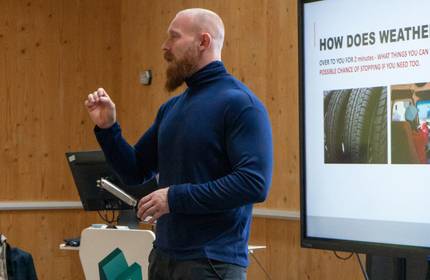 A man with a shaved head and beard stands in front of a large display screen giving a presentation.