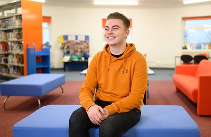A person wearing an orange hoodie sits smiling on a blue bench in a brightly lit room. The space has colourful furniture, with shelves of books and a bulletin board visible in the background.