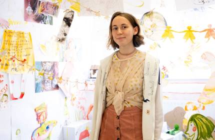 A young woman stands in front of a wall covered with colourful drawings and art. She is wearing a light-coloured jacket, a patterned top, and a coral skirt. The art features various abstract and playful designs.